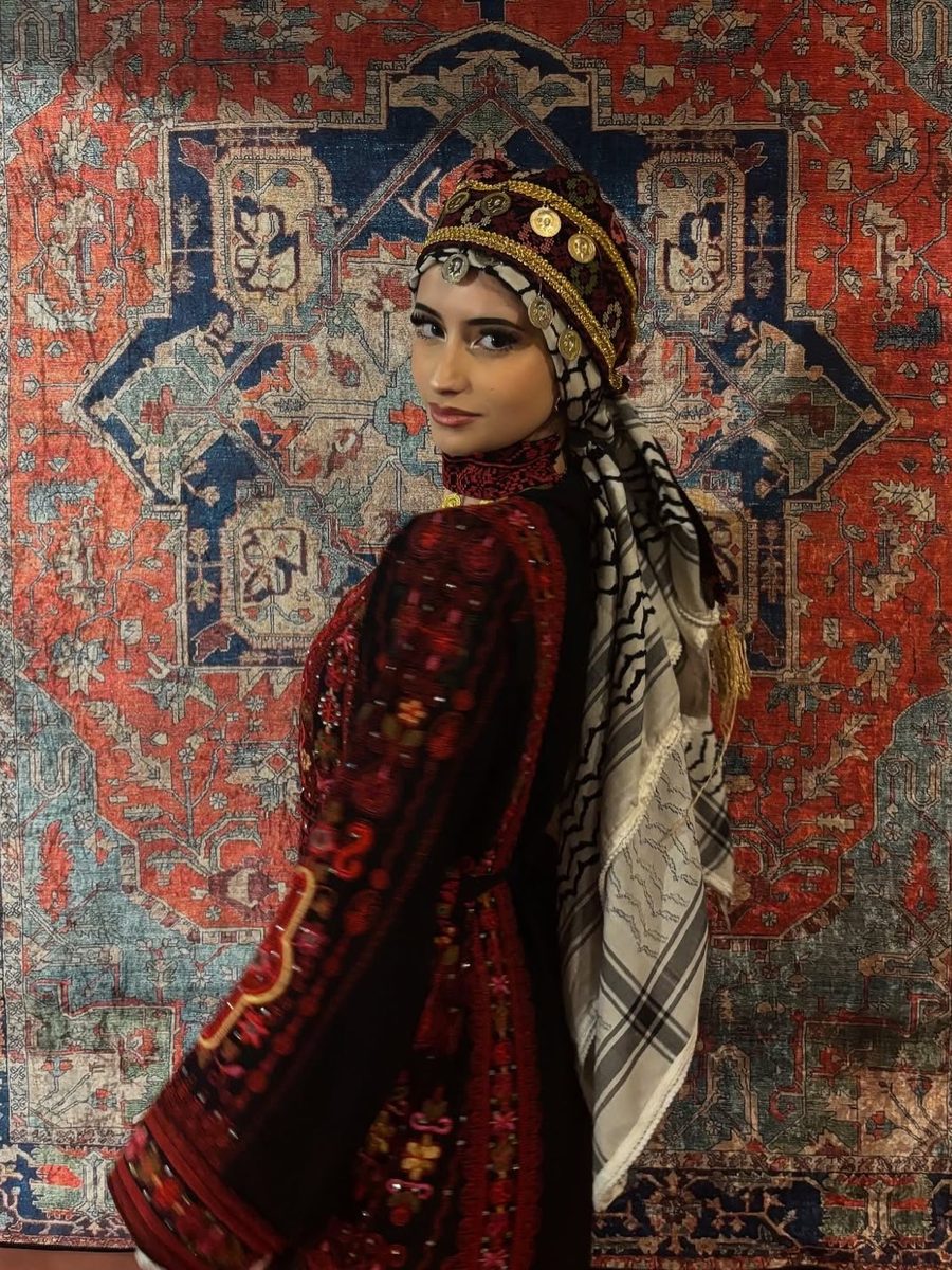 Aisha looking over her shoulder in a red embroidered dress, coin headpiece and patterned scarf, in front of a woven rug.