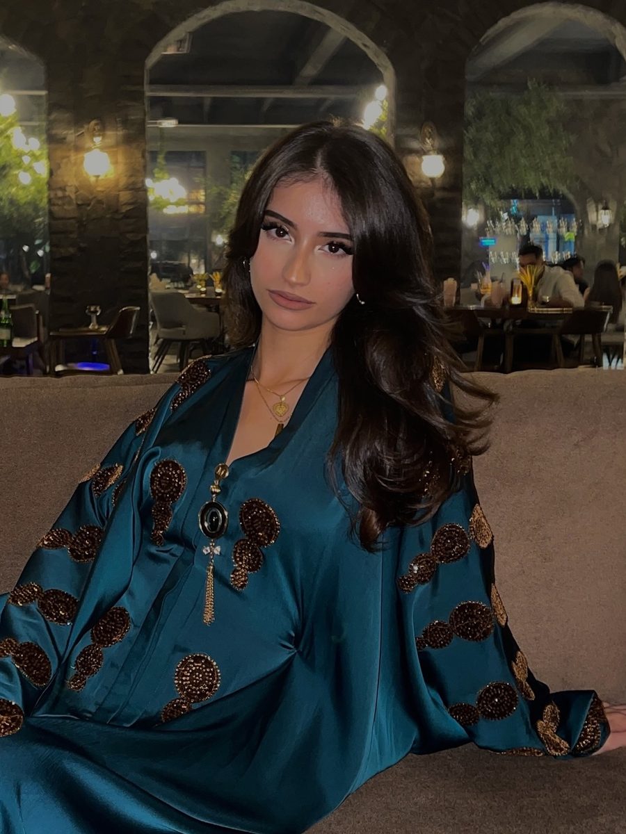 Aisha seated on a banquette in a teal satin kaftan with bronze embroidery, in a lit restaurant.